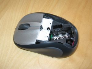 A wireless computer mouse. Part of the cover has been removed. There's no battery, but the space for a AA battery is clearly labeled, with an outline to show that the positive end of the battery should go to the right.
